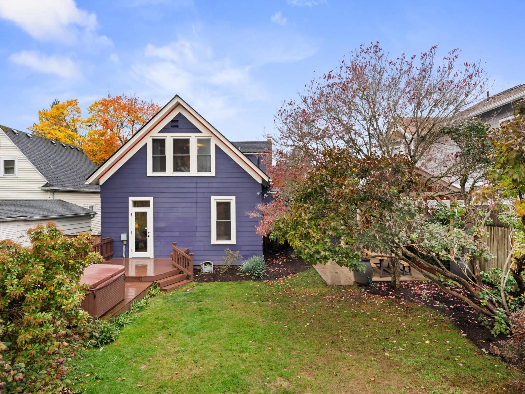 A quaint purple house with white trim and an A-frame roof sits in a backyard surrounded by autumn trees. A wooden deck with a hot tub is visible on the left, and a small lawn and garden area add to the cozy setting.