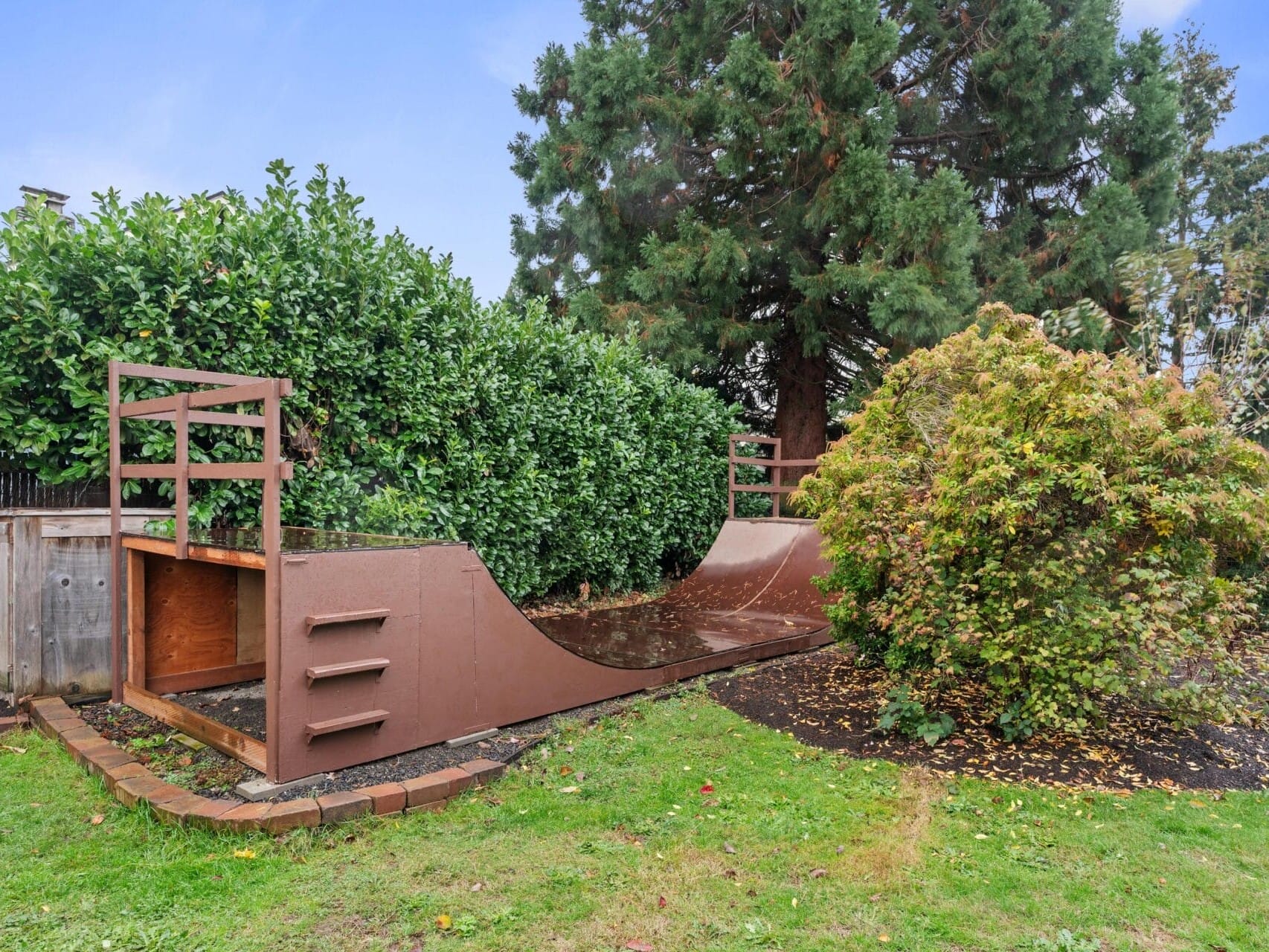 A wooden half-pipe skateboard ramp is set up in a backyard surrounded by lush greenery, including a large tree and bushes. The grass is green and slightly wet, suggesting recent rain.