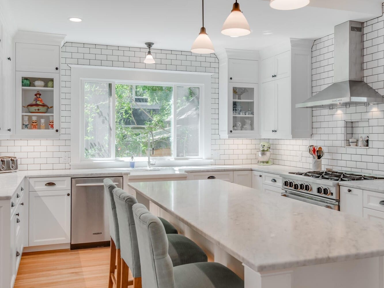 A bright, modern kitchen with white cabinets and subway tile backsplash. It features a large marble island with three upholstered stools, stainless steel appliances, and pendant lights. A window provides natural light and a view of greenery outside.