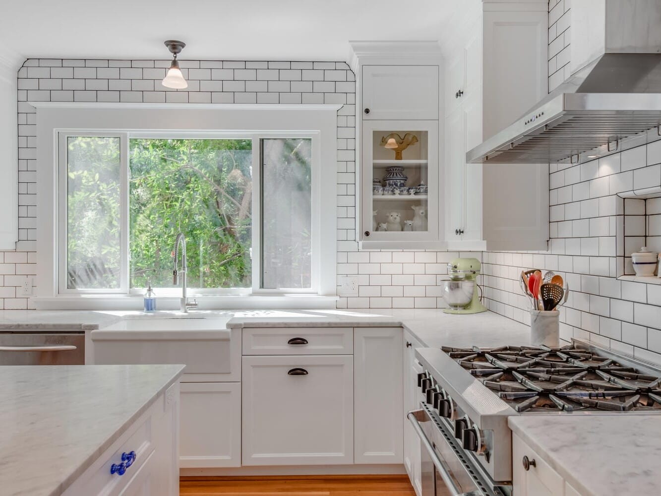 A bright kitchen with white cabinets, a marble island, and a stainless steel stove. Large window over the sink offers a view of greenery outside. The backsplash is made of white subway tiles, and various kitchen utensils are on the counters.