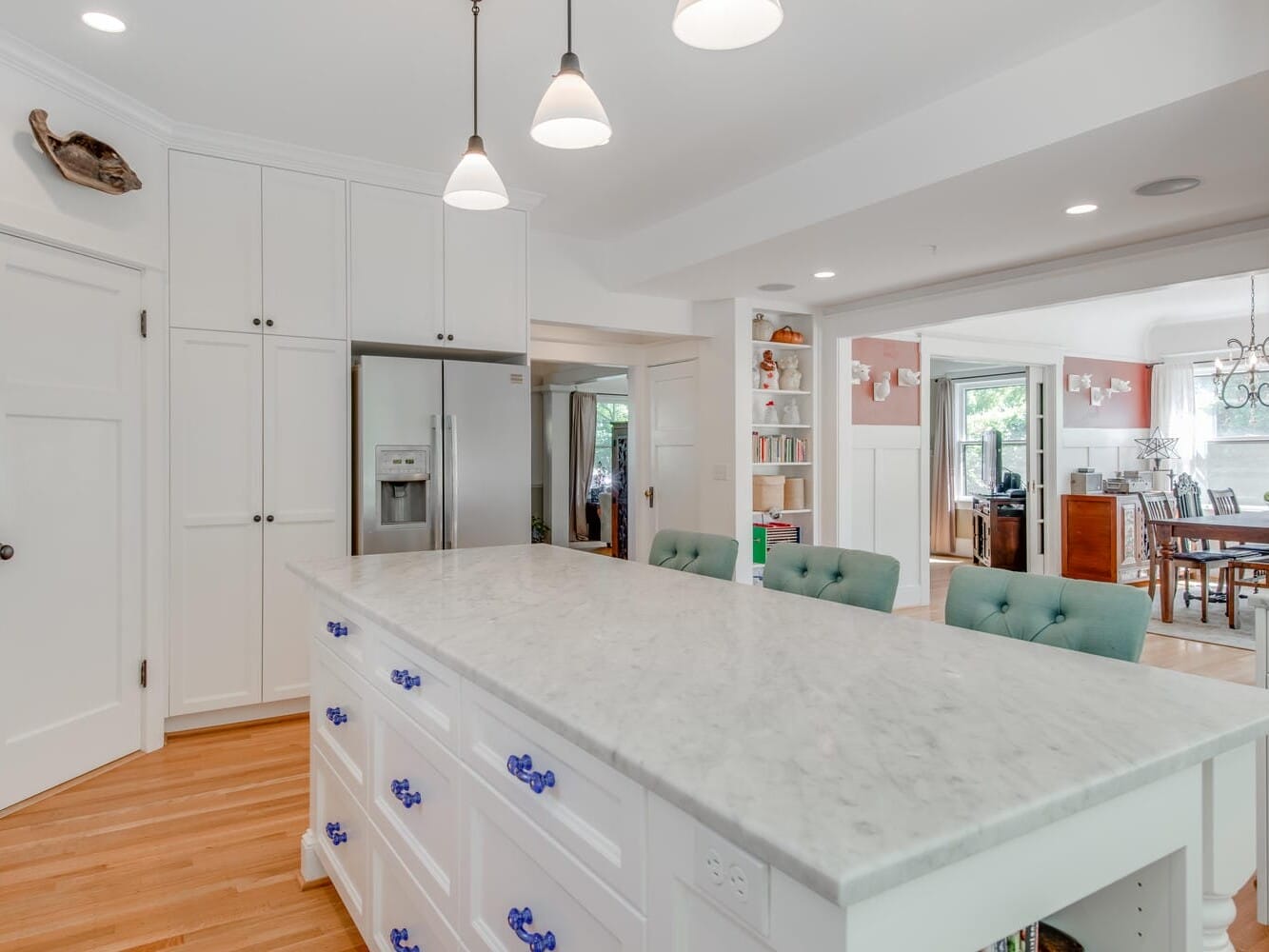 A bright kitchen with a marble island features white cabinets, stainless steel appliances, and teal barstools. Pendant lights hang above the island. The adjacent dining area is visible through an open doorway, showcasing a wooden table and chairs.