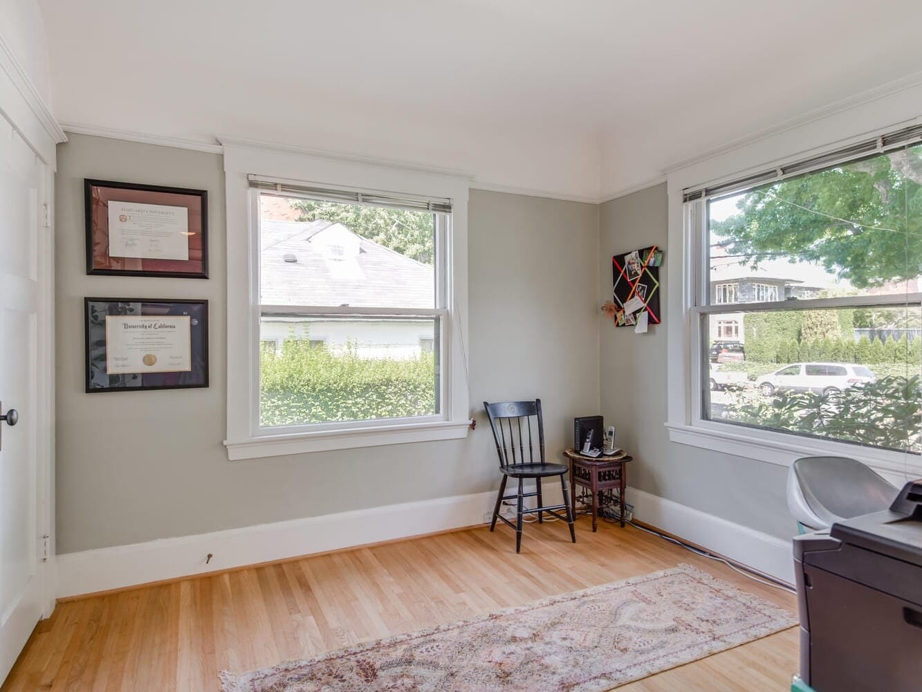 A bright room with two large windows, a wooden floor, and light gray walls. A black chair and small table with a camera are near the window. Framed certificates hang on the wall, and a colorful geometric artwork is displayed nearby.