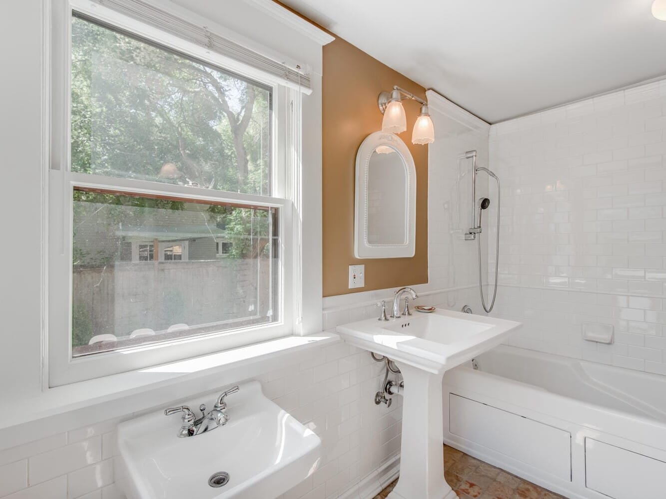 A bright bathroom with a window, white fixtures, and tile. It features a pedestal sink, a wall mirror, a bathtub with a shower, and beige walls. A small window offers a view of trees and a neighboring house.