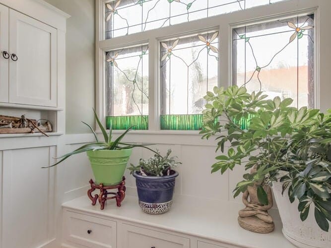 A cozy interior space with a stained glass window, featuring green geometric accents. Below is a white bench with built-in storage, adorned with three potted plants. A decorative rug lies on the wooden floor.