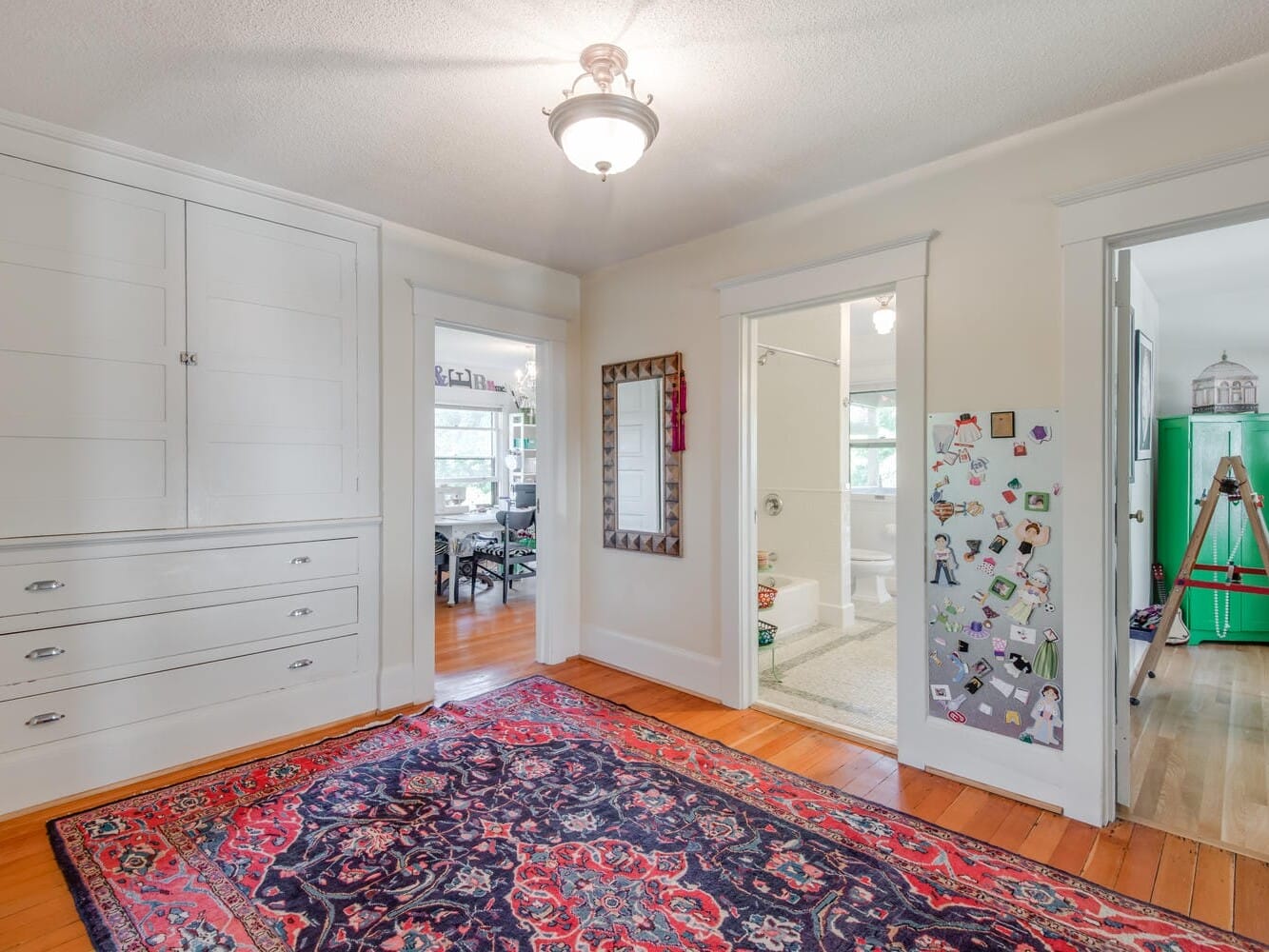 A hallway with a colorful patterned rug on hardwood floors. There are built-in cabinets on the left, a large mirror in the center, and three open doorways leading to various rooms. A telescope is set up by the window in the right room.