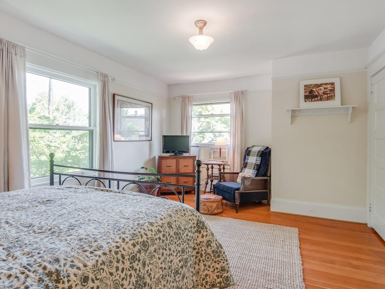 A bright bedroom with a wrought iron bed, covered in a green and white floral quilt. Two large windows allow natural light. Theres a wooden floor, a small TV on a stand, an armchair with a plaid blanket, and a framed art piece on the wall.