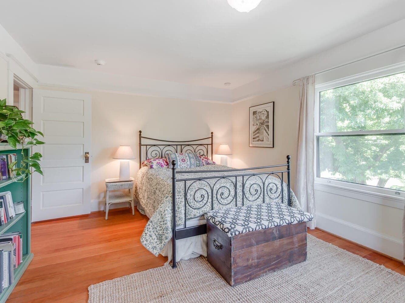 A cozy bedroom with a wrought iron bed adorned with patterned bedding. Two bedside lamps are lit, and a wooden chest sits at the foot of the bed. A large window with curtains offers a view of greenery. A bookshelf and a plant are on the left.