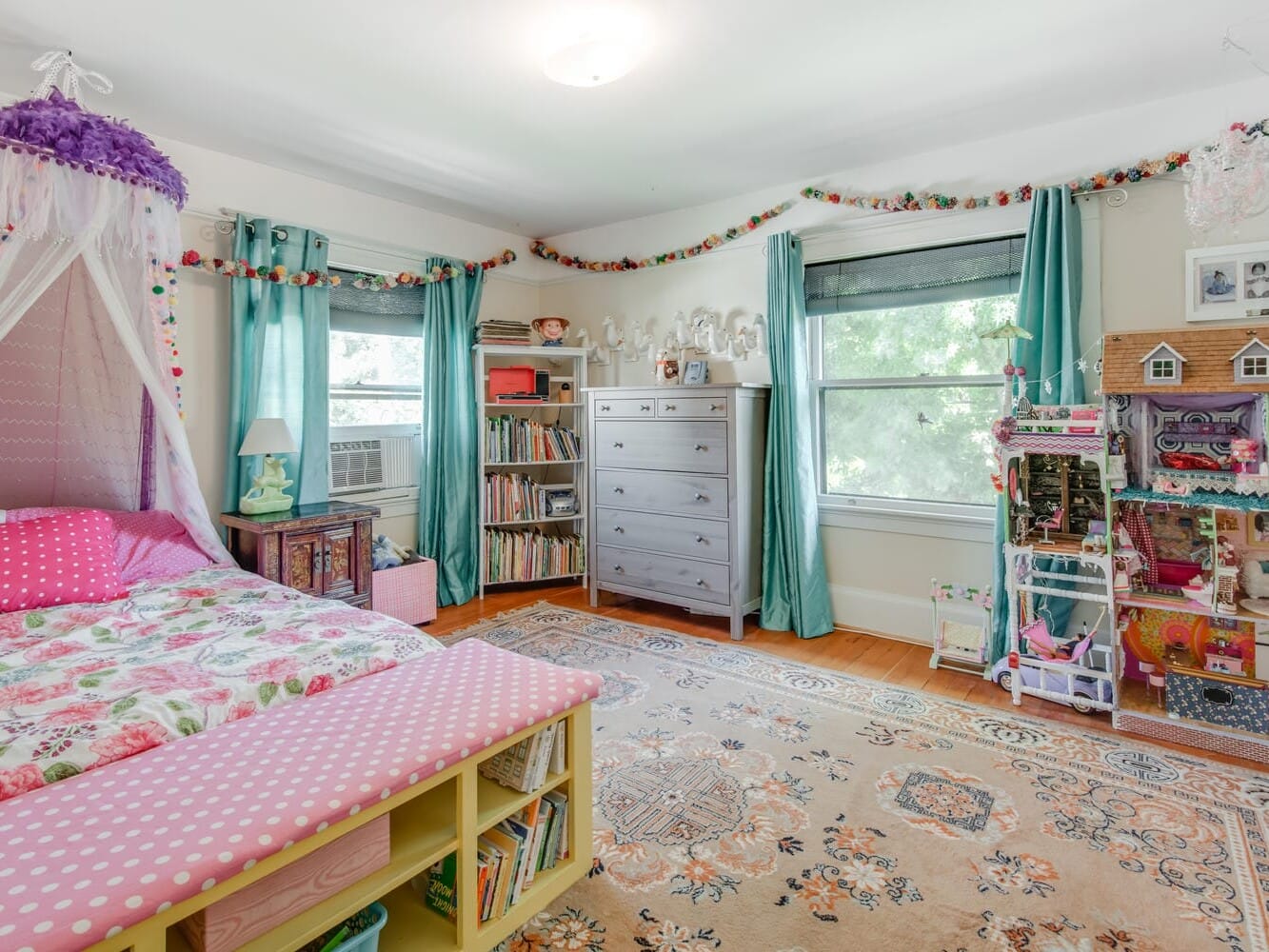 A bright, colorful childrens bedroom with a floral bed, patterned rug, and canopied corner. Shelves full of books and a large dollhouse are in view. Blue curtains hang by large windows, and a pink bench sits at the foot of the bed.