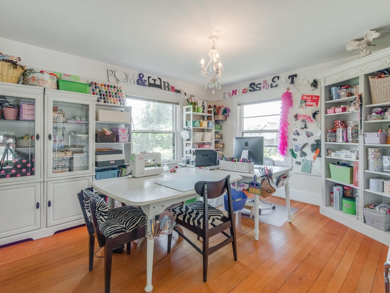 A brightly lit craft room with a chandelier, featuring tables and various chairs. Surrounding shelves are filled with craft supplies, sewing essentials, and decorative items. A computer sits on one of the tables near a window with natural light.