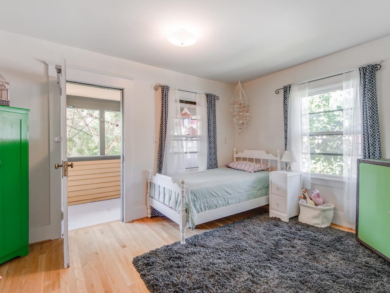 A bright, airy bedroom features a white bed with light green bedding, a soft gray rug, and two windows with blue curtains. A green wardrobe and a small cabinet add pops of color. The door is open, showing a glimpse of a sunlit balcony.