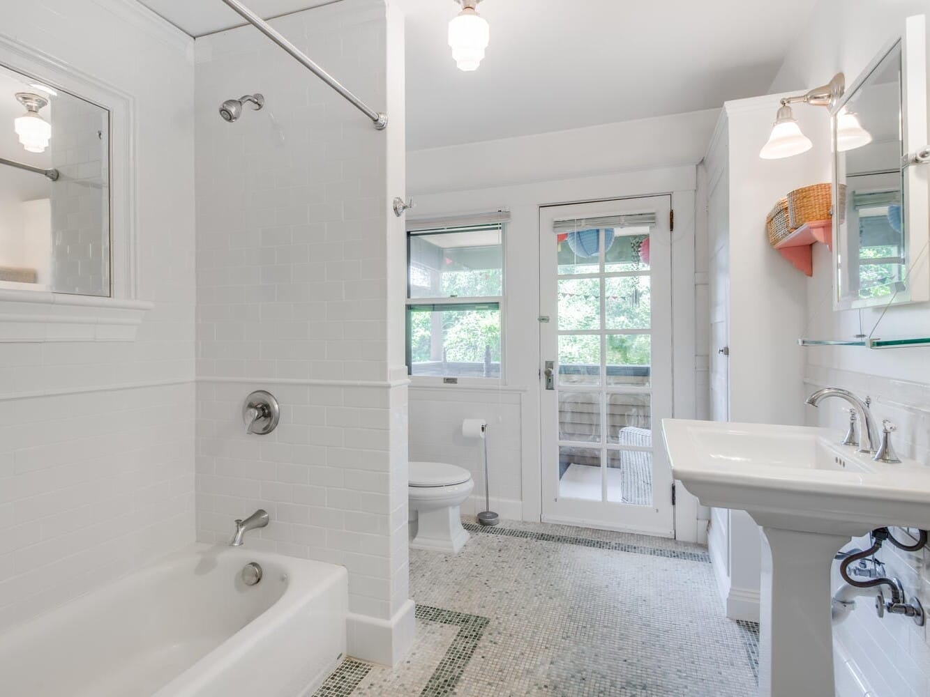 Bright bathroom with white tiles, featuring a bathtub, shower, pedestal sink, and toilet. A door with glass panels leads outside, and theres a small window above the toilet. The floor is tiled, and there are wall-mounted lights.