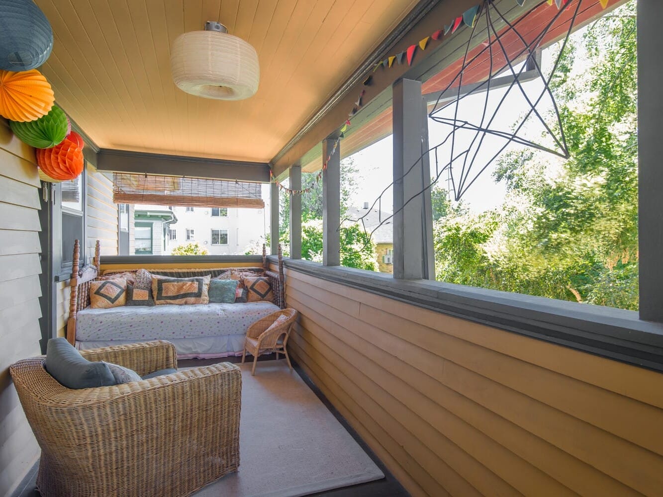 A cozy porch with a wicker chair and a cushioned bench adorned with pillows. The space features colorful paper lanterns, a hanging lamp, and decorative lights, with greenery visible outside through the open railing.