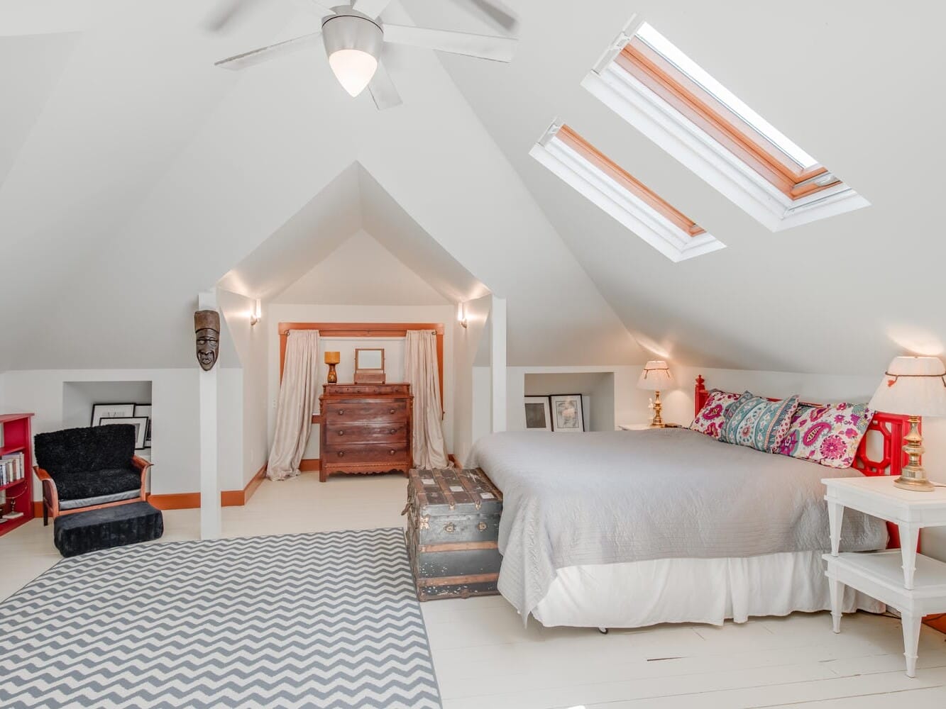 A cozy attic bedroom with sloped ceilings. It features a double bed with colorful pillows, a zigzag rug, a vintage chest, skylights, a ceiling fan, bookshelves, and a small armchair. Warm lighting adds to the inviting atmosphere.