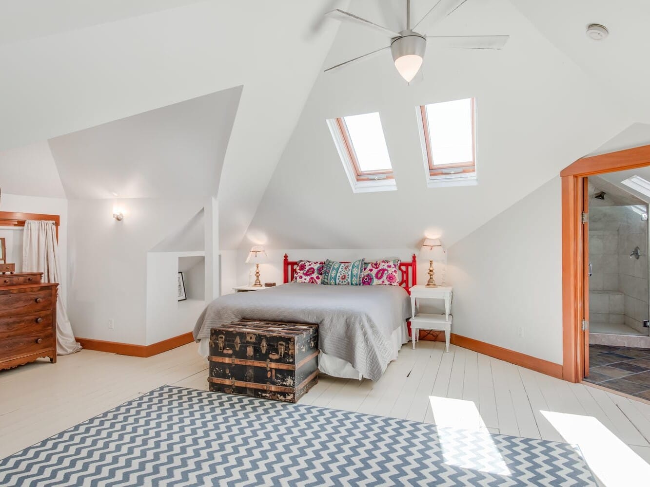 A bright, airy attic bedroom with sloped ceilings and two skylights. The room features a double bed with colorful pillows, a wooden chest at the foot, a dresser, and access to a bathroom. A patterned rug covers part of the wooden floor.