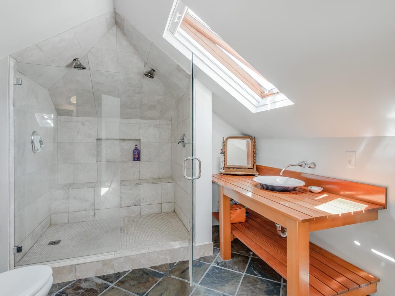 A modern bathroom with a sloped ceiling featuring a glass-enclosed shower with dual showerheads and beige tiles. A wooden vanity with a vessel sink and a skylight above illuminates the space, and the floor has dark slate tiles.