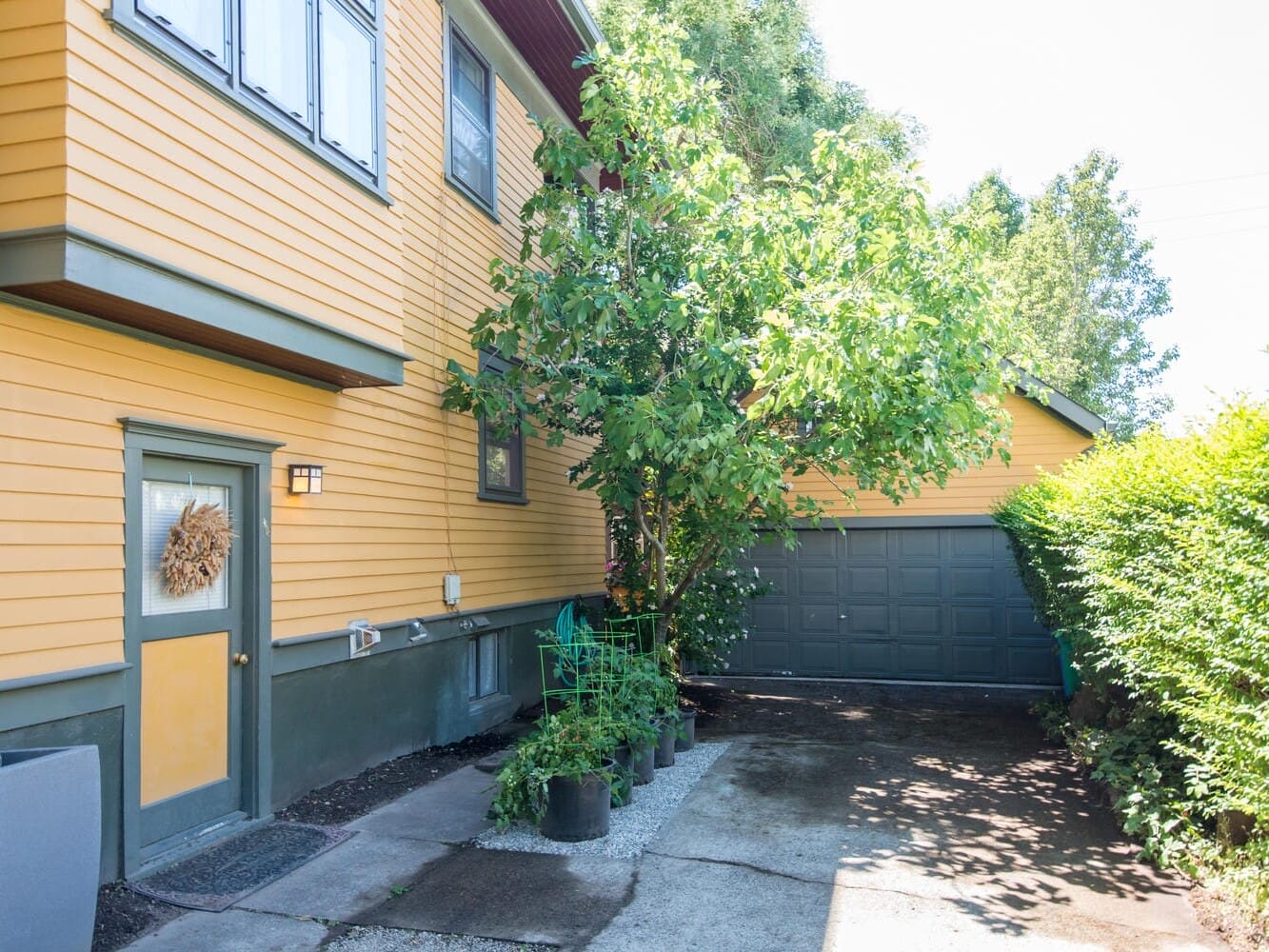 A yellow house with blue trim, a wreath on the door, and a small tree in a pot near the entrance. A driveway leads to a gray garage surrounded by green bushes and trees.