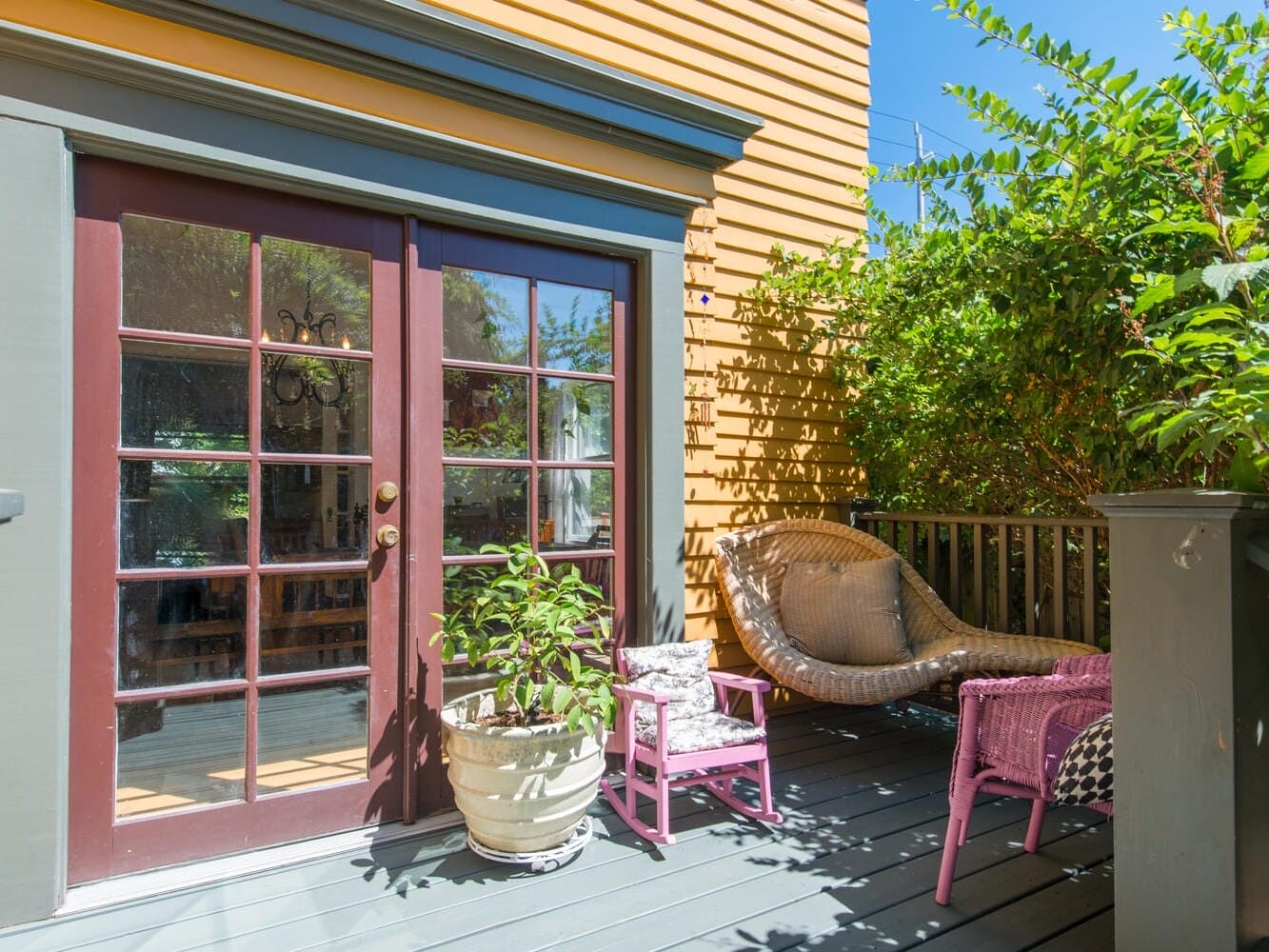 A sunny porch with a cozy seating area. Theres a rattan chair and a pink side table on a gray deck. A potted plant sits near the door, which has large glass panes. Lush greenery surrounds the porch, creating a peaceful outdoor retreat.