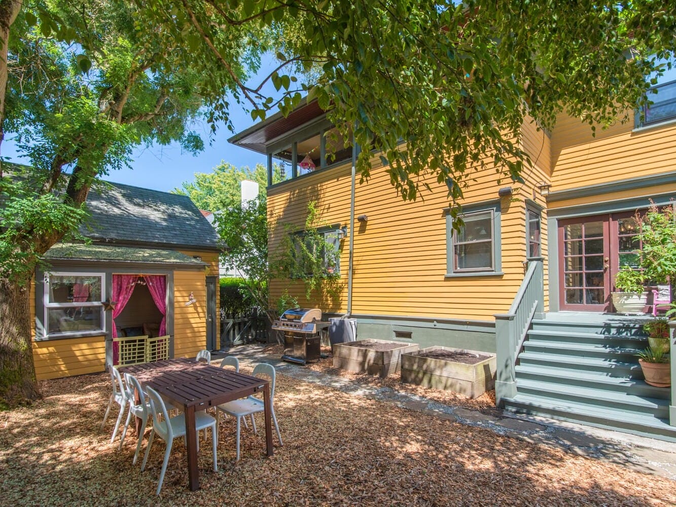 A backyard scene with a yellow house and matching shed. Theres a wooden table with chairs on a ground covered in wood chips. A barbecue grill is nearby. Trees provide shade, and a staircase leads up to a door on the house.