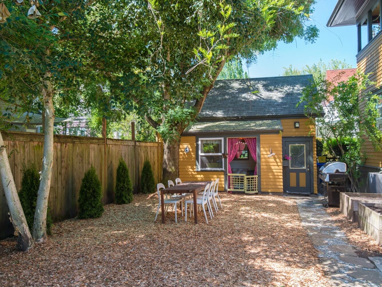 A cozy backyard with a wooden table and chairs on mulch. Surrounded by trees and shrubs, theres a quaint yellow house with pink curtains, a BBQ grill to the right, and a wooden fence lining the yard.