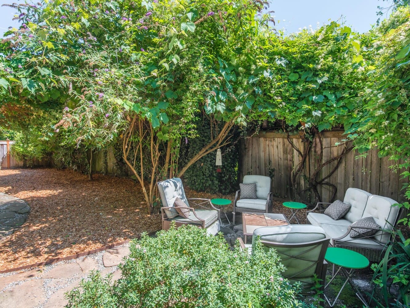 A lush backyard garden with cushioned patio chairs and small green tables arranged on a stone-paved area. Surrounded by dense greenery and tall bushes, the space provides a tranquil outdoor setting.