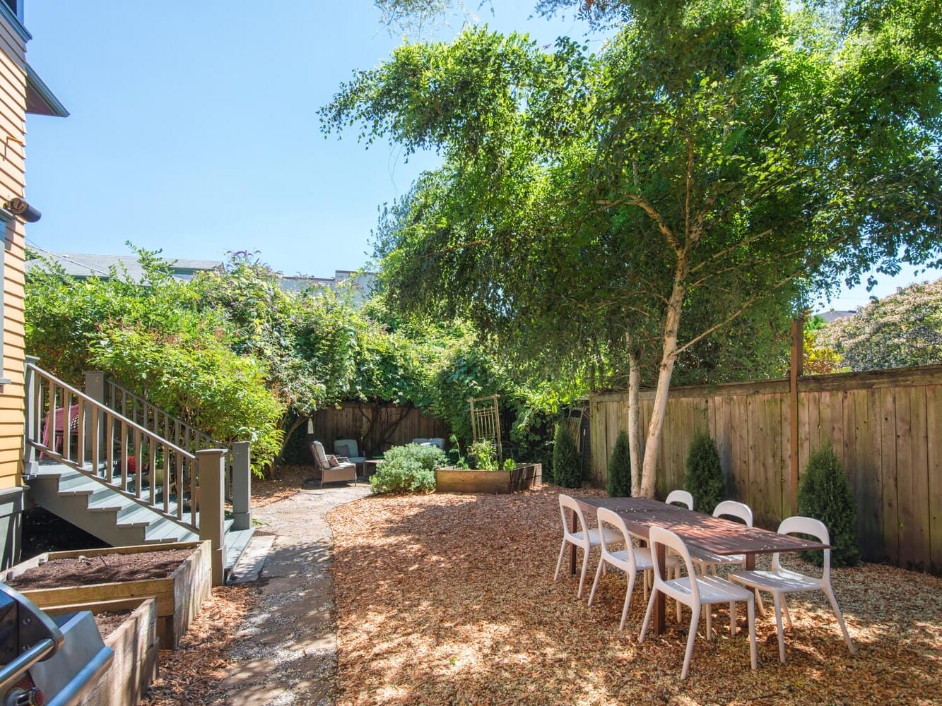 A sunny backyard with woodchip ground cover features a wooden table and white chairs. A barbecue grill is in the foreground, and a garden with lush trees and shrubs adds greenery around the fenced area. A set of steps leads up to a house.