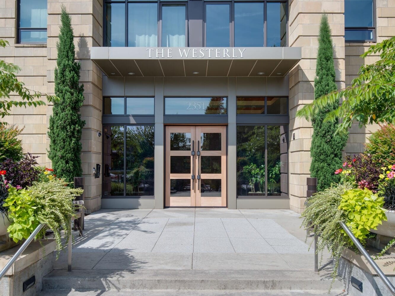 Entrance of The Westerly building featuring glass doors and large windows, flanked by tall green plants. Two symmetrical planters with lush greenery and red flowers are placed on either side of the walkway leading to the entrance.