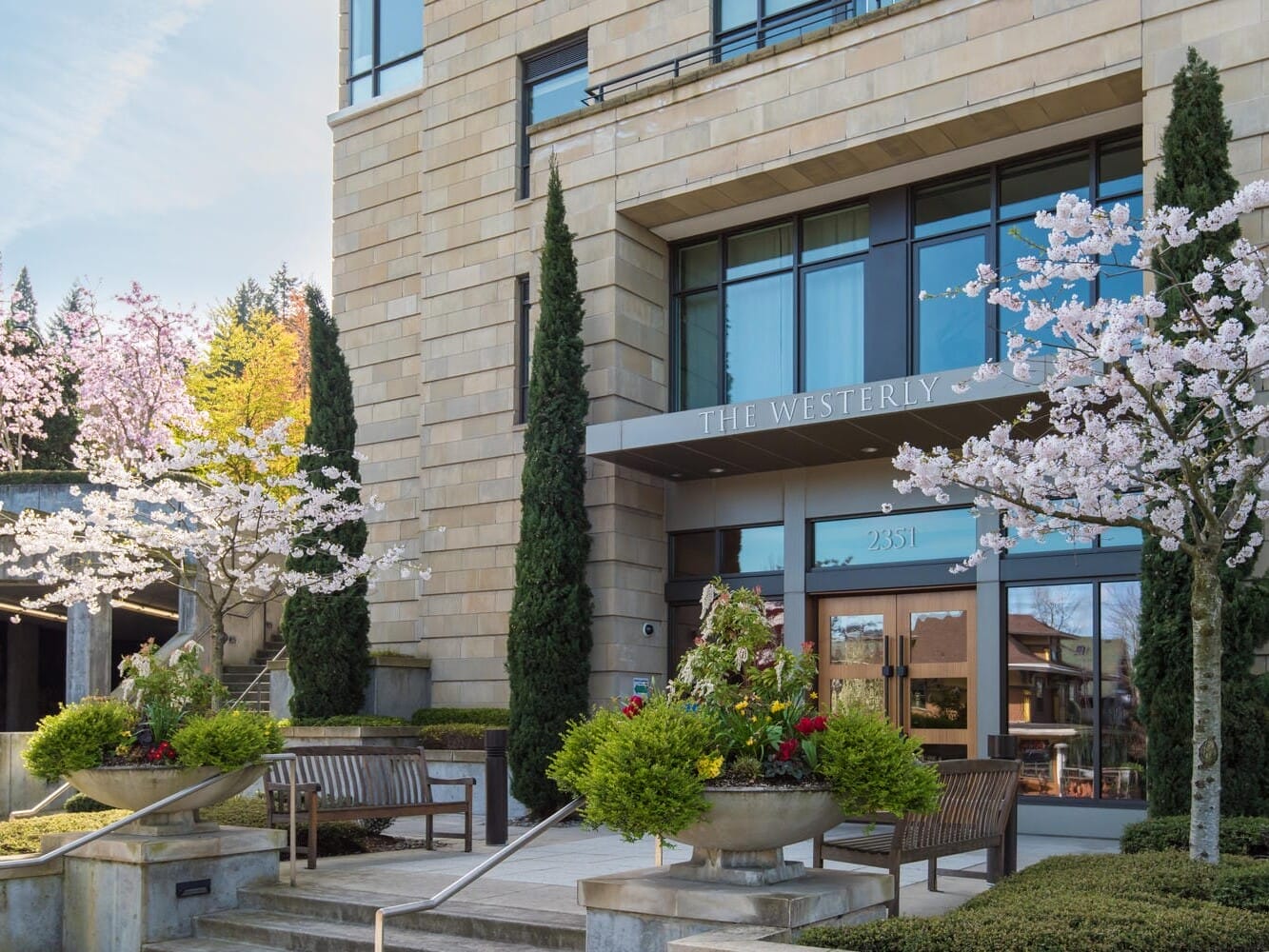 The image shows the facade of a building labeled The Westerly, featuring a stone exterior with large windows. There are blooming cherry blossom trees and neatly arranged planters and benches surrounding the entrance.