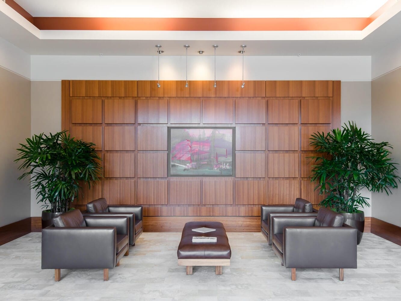 A modern, well-lit waiting area with four leather chairs and a central ottoman. The room features a wooden paneled wall with a framed artwork, flanked by two large potted plants on either side.