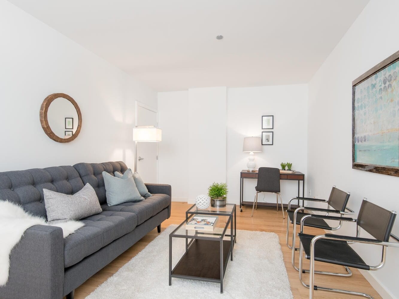 A modern living room with a gray sofa, a round mirror on the wall, a floor lamp, and a glass-top coffee table on a white rug. A small desk with a lamp and chair is against the back wall, and two black chairs face the sofa.