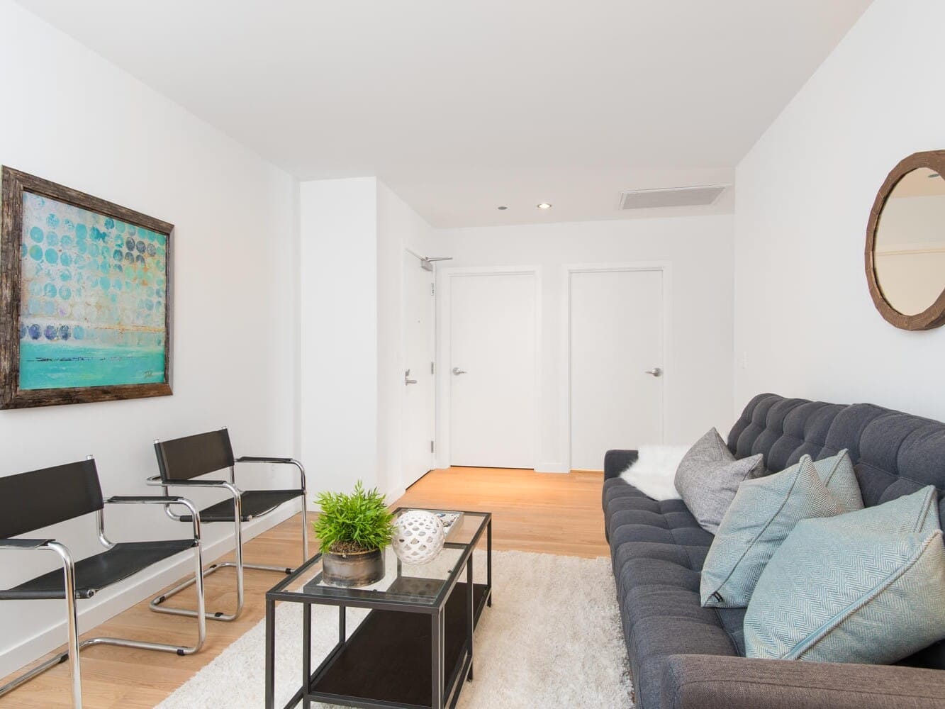 A modern living room with a gray sofa, blue and gray cushions, two black chairs, and a glass coffee table with a plant. Theres a framed abstract painting on the wall and a round mirror. The room has wooden floors and white walls.