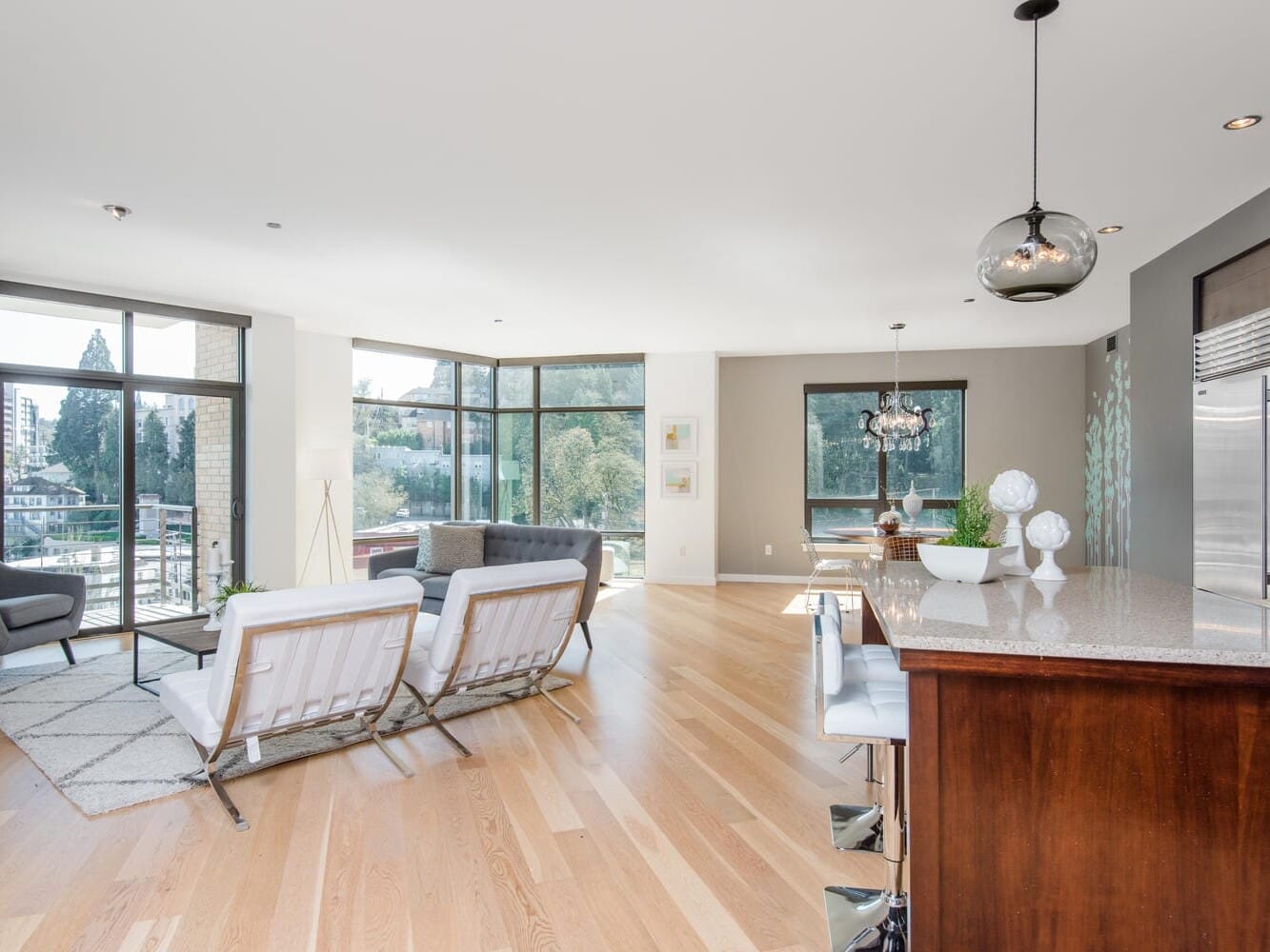 A modern, open-concept living space with large windows showcasing a city view. The room features light wood flooring, a kitchen island with bar stools, contemporary furniture, and a mix of neutral and dark accents. Natural light fills the area.