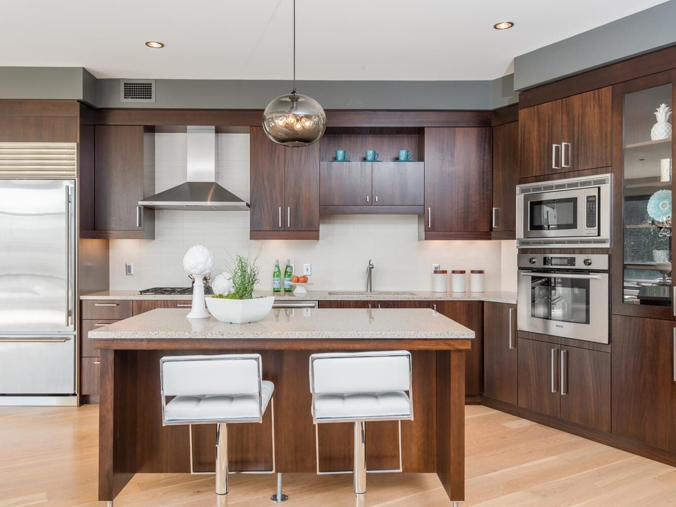 Modern kitchen with dark wood cabinets, stainless steel appliances, and a central island with two white bar stools. A pendant light hangs above the island, and decorative items are placed on the counter. Light wood flooring complements the space.