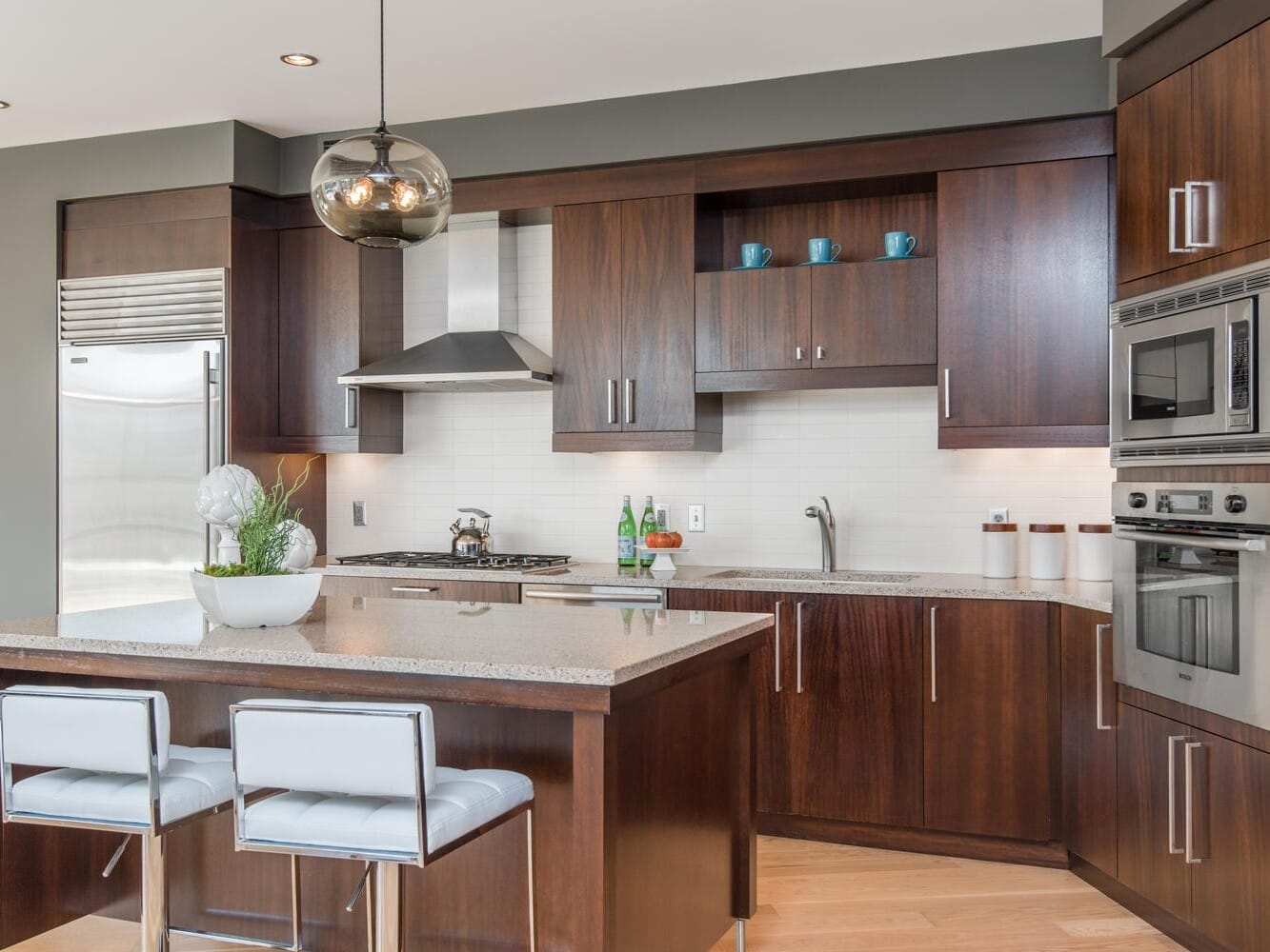 Modern kitchen with wooden cabinets and stainless steel appliances. An island with a granite countertop has two white cushioned stools. A bowl with a plant decorates the island, and a pendant light hangs above.