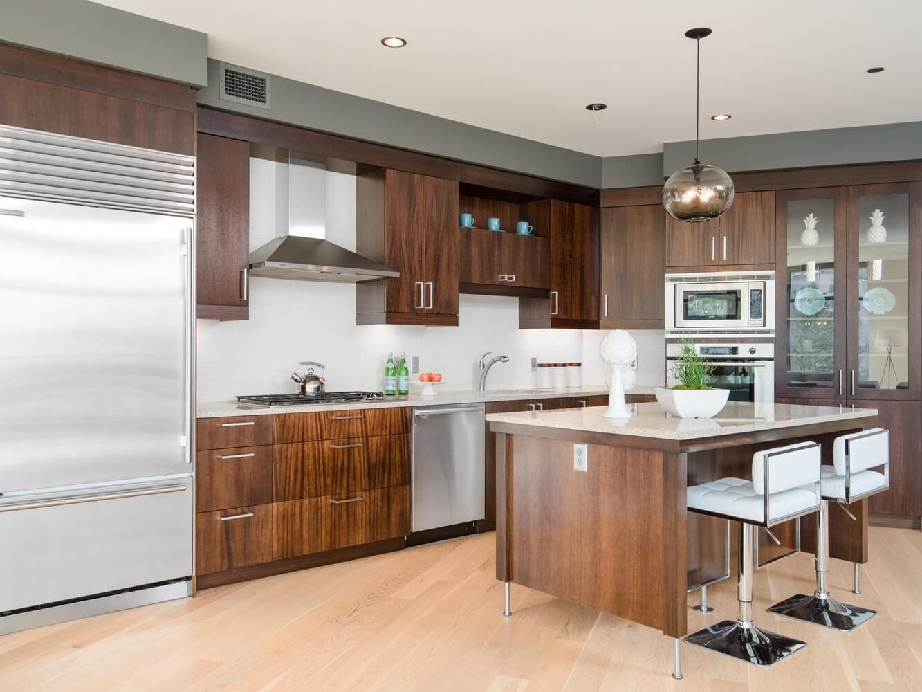 Modern kitchen with dark wood cabinetry, stainless steel appliances, and a central island with bar stools. A pendant light hangs above the island, and the room features a mix of open shelving and glass-front cabinets.