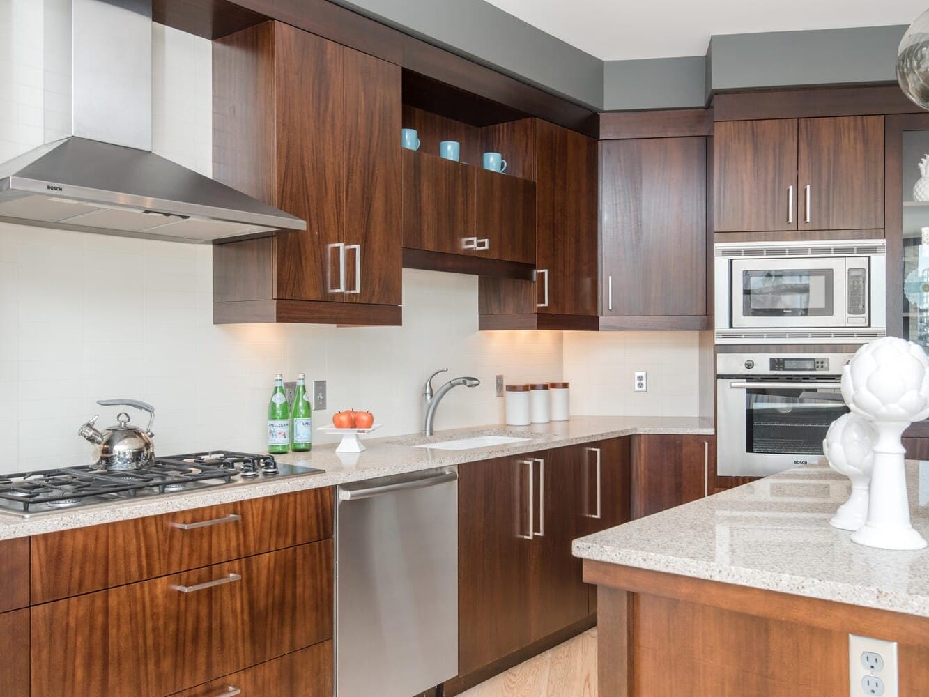 Modern kitchen with dark wood cabinetry, stainless steel appliances, and granite countertops. A silver range hood and a teapot sit on the stove. Decorative vases and fruit are on the counter, with a view of the city visible through a window.