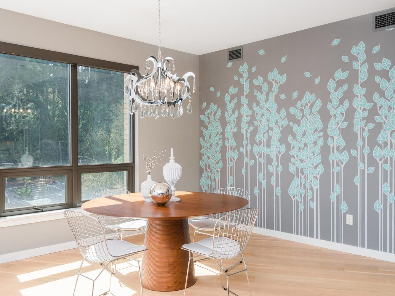 A modern dining area featuring a round wooden table with three white wire chairs. A decorative chandelier hangs above. The wall displays a leaf pattern mural in shades of light blue and gray. Large window illuminates the space with natural light.