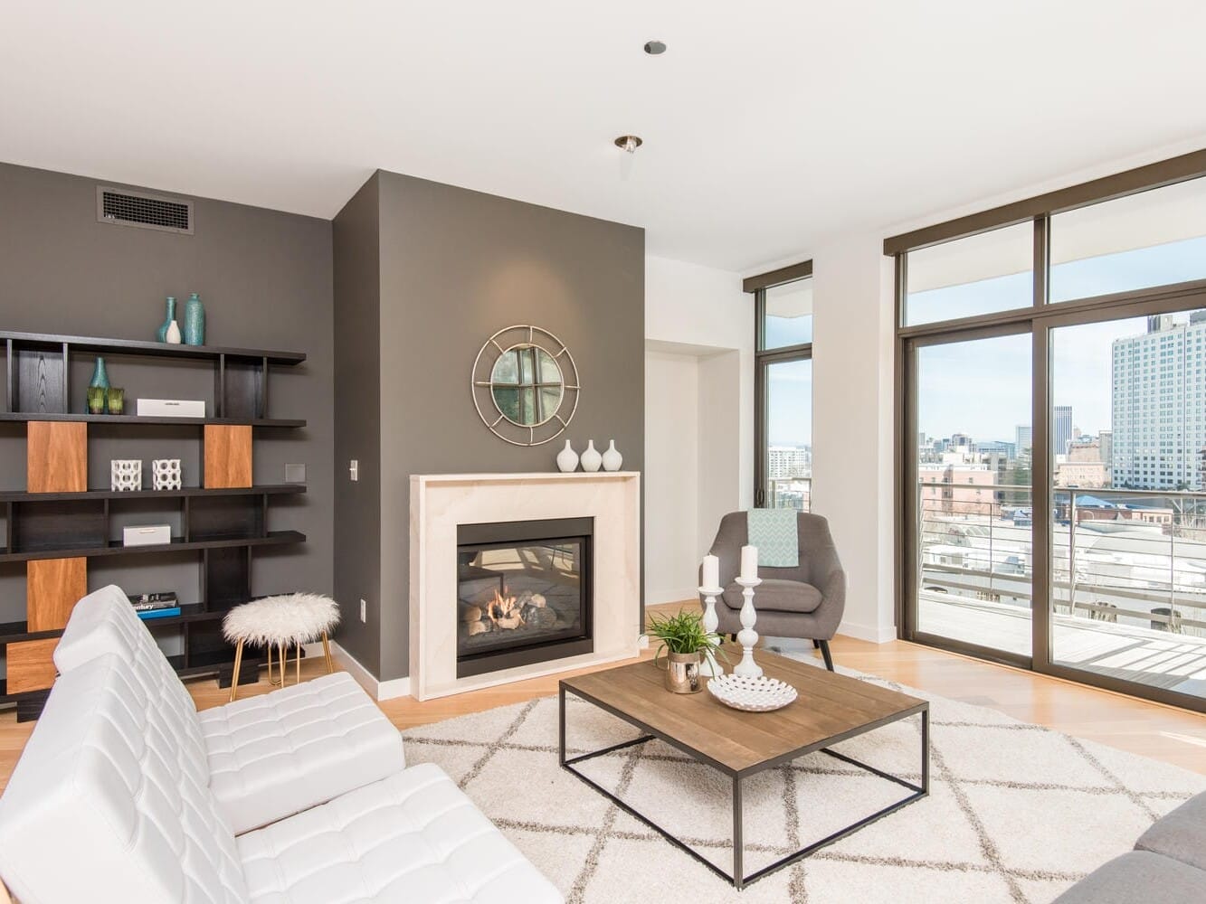 A modern living room with a white sofa, gray armchair, and a wooden coffee table. Features a fireplace with a round mirror above it, bookshelves with decor, large windows, and a city view. Neutral tones and minimalistic design.