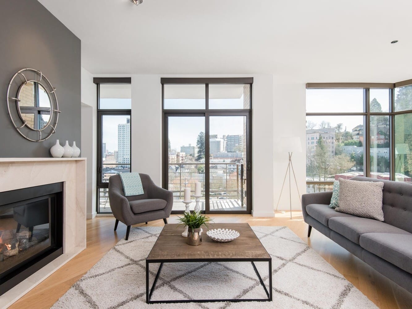 A modern living room with large windows offering a city view. It features a gray armchair, a gray sofa with patterned pillows, a wooden coffee table, a decorative fireplace, and a circular mirror above it. The room is bright and spacious.