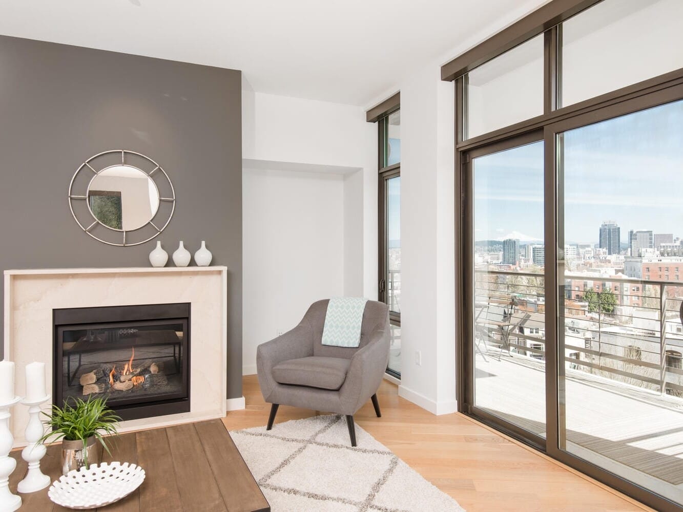 A modern living room with a gray armchair, a fireplace with a round mirror above it, and a wooden coffee table. Large windows reveal a balcony and a cityscape view. The decor is minimal with neutral tones.