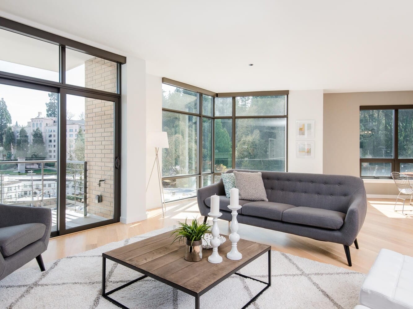 A modern living room with large windows, a gray sofa and armchair, a coffee table with candles and a plant, and a view of trees and buildings outside. The room features a light carpet and a dining area in the background.