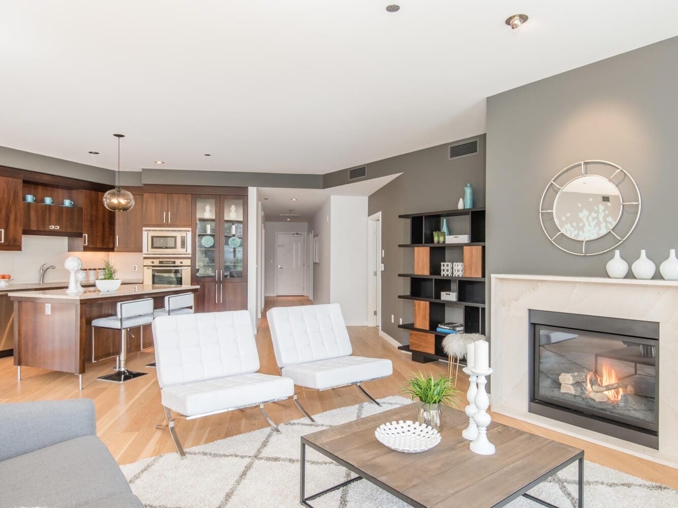 Modern open-concept living room and kitchen featuring white chairs, a gray couch, a contemporary fireplace, dark wood cabinetry, and sleek stainless steel appliances. The space is decorated with neutral tones and minimalist accessories.