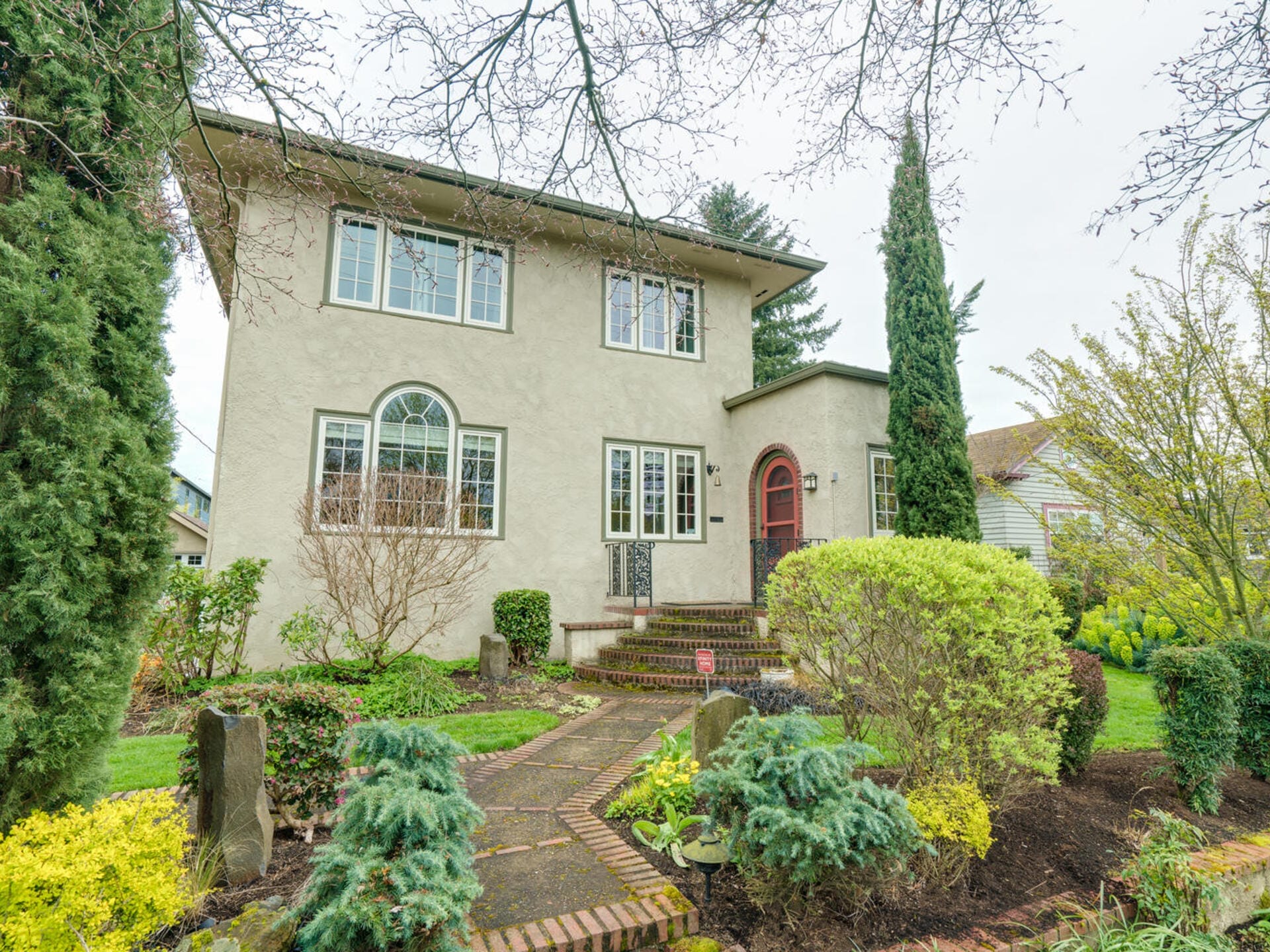 A charming two-story stucco house with large windows and a red front door, surrounded by a lush garden. The garden features neatly trimmed shrubs, colorful plants, and a stone pathway leading to the entrance. Trees frame the scene.