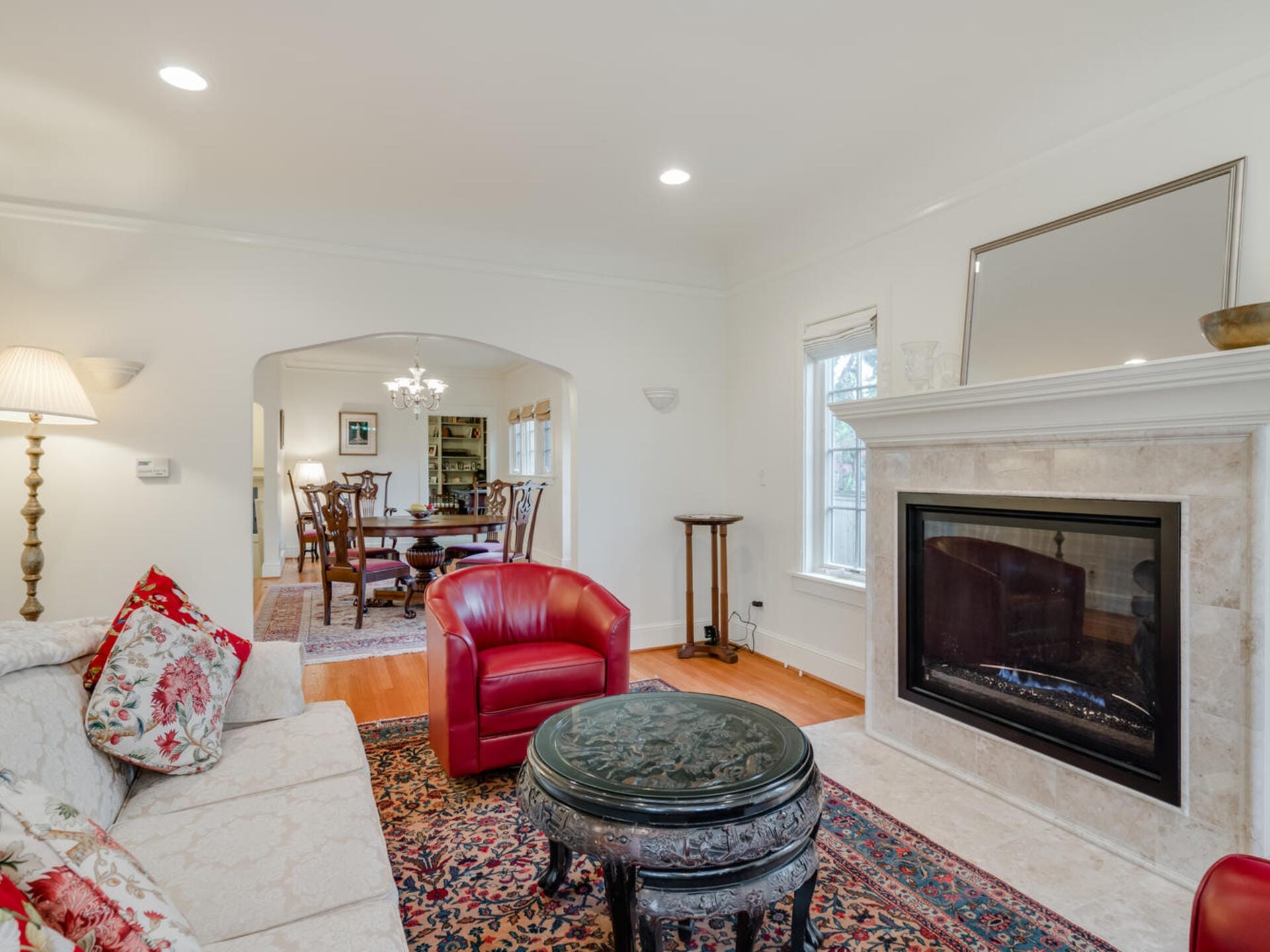 A cozy living room with white walls features a red armchair, a floral-patterned sofa, and a dark ornate coffee table on a colorful rug. A fireplace is built into the wall, and a dining area with a wooden table and chairs is visible through an archway.