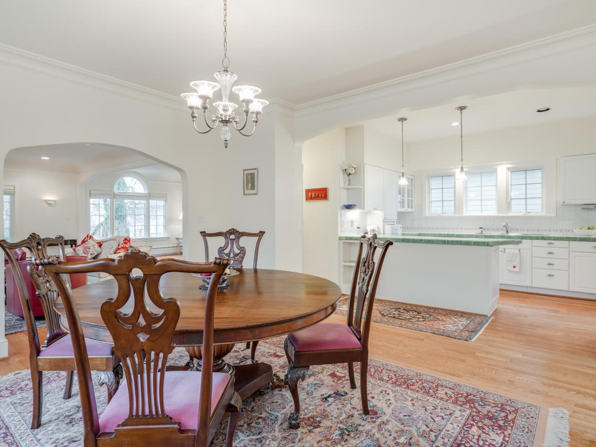 A dining room with a wooden table and six chairs on a patterned rug. Theres an arched entryway leading to a sitting area with a window. The room opens into a bright kitchen with a large island and pendant lights, all with light-colored walls and wooden floors.