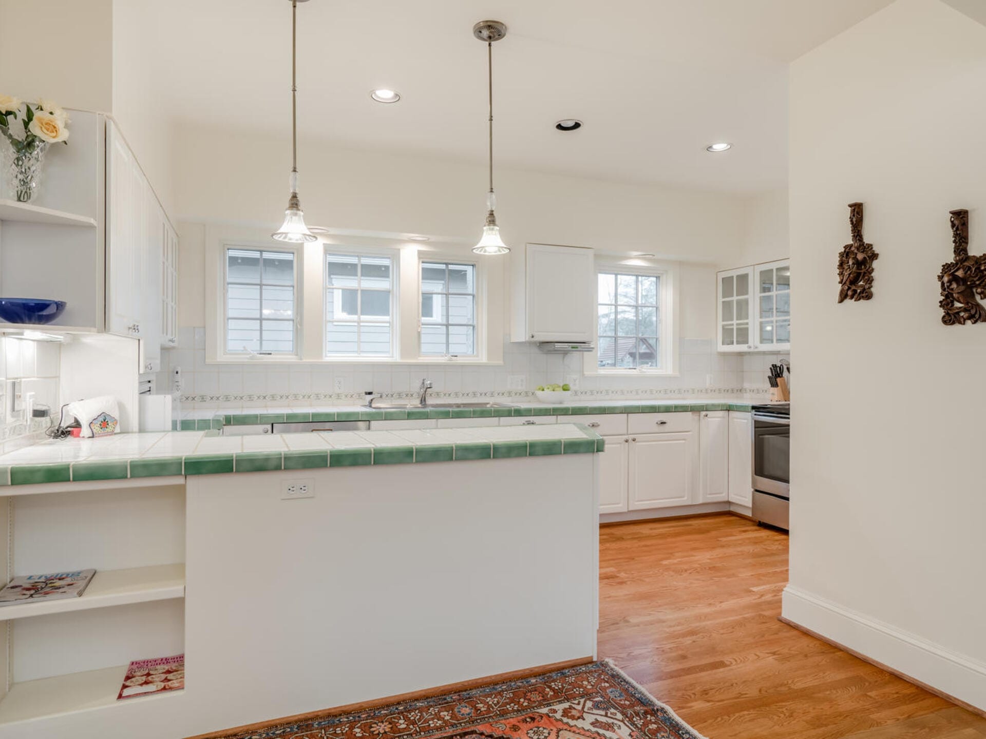 A bright kitchen with white cabinets, a green-tiled countertop, and stainless steel appliances. Pendant lights hang above the counter. Windows line the wall, and a patterned rug lies on the wooden floor. Decorative wall hangings are visible.