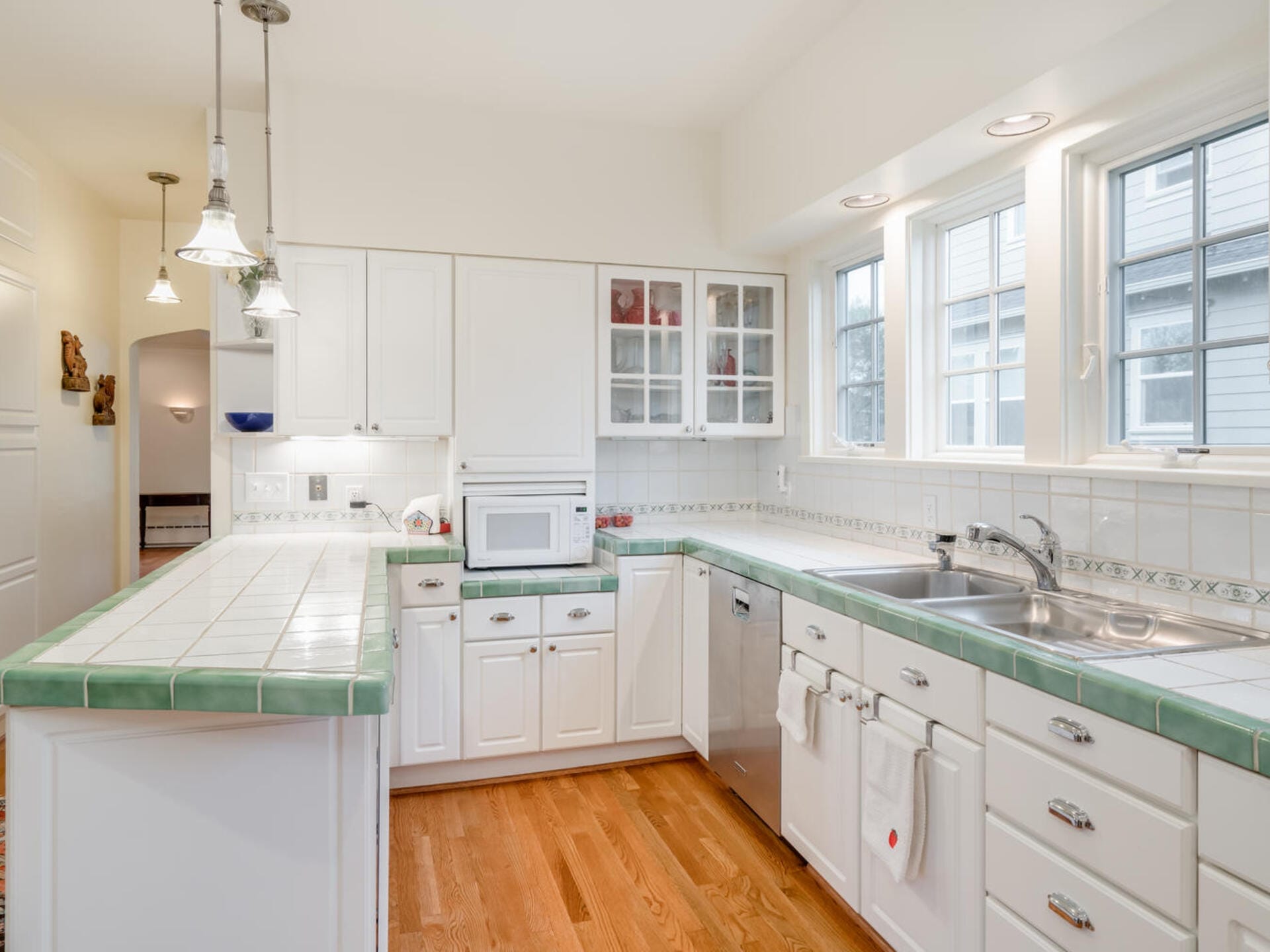 A bright kitchen with white cabinets and green-tiled countertops. It features a stainless steel dishwasher, microwave, and double sink. Three pendant lights hang over the island, and natural light streams in through a row of windows above the sink.
