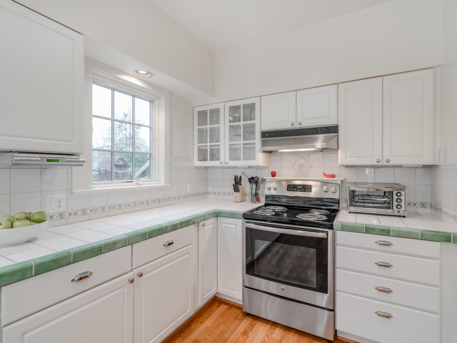 A bright kitchen with white cabinets and green-tiled countertops. It features an electric stove, a toaster oven, and utensils in a holder next to the stove. A window above the counter lets in natural light. A fruit bowl is on the left.