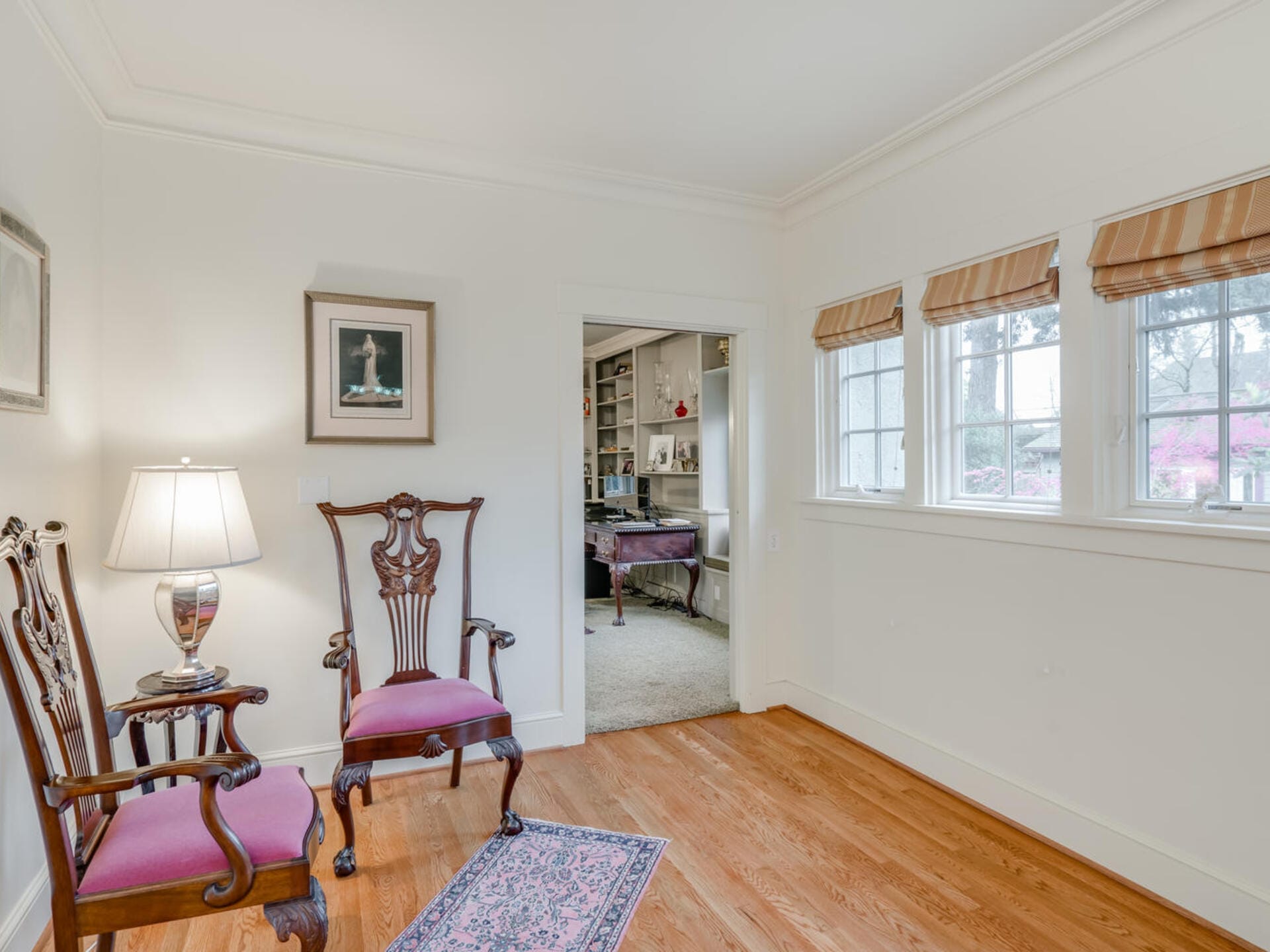 A cozy room with two ornate wooden chairs with pink cushions, a small table with a lamp, and a framed picture on the wall. A doorway leads to another room with bookshelves. Three windows with striped shades let in natural light.