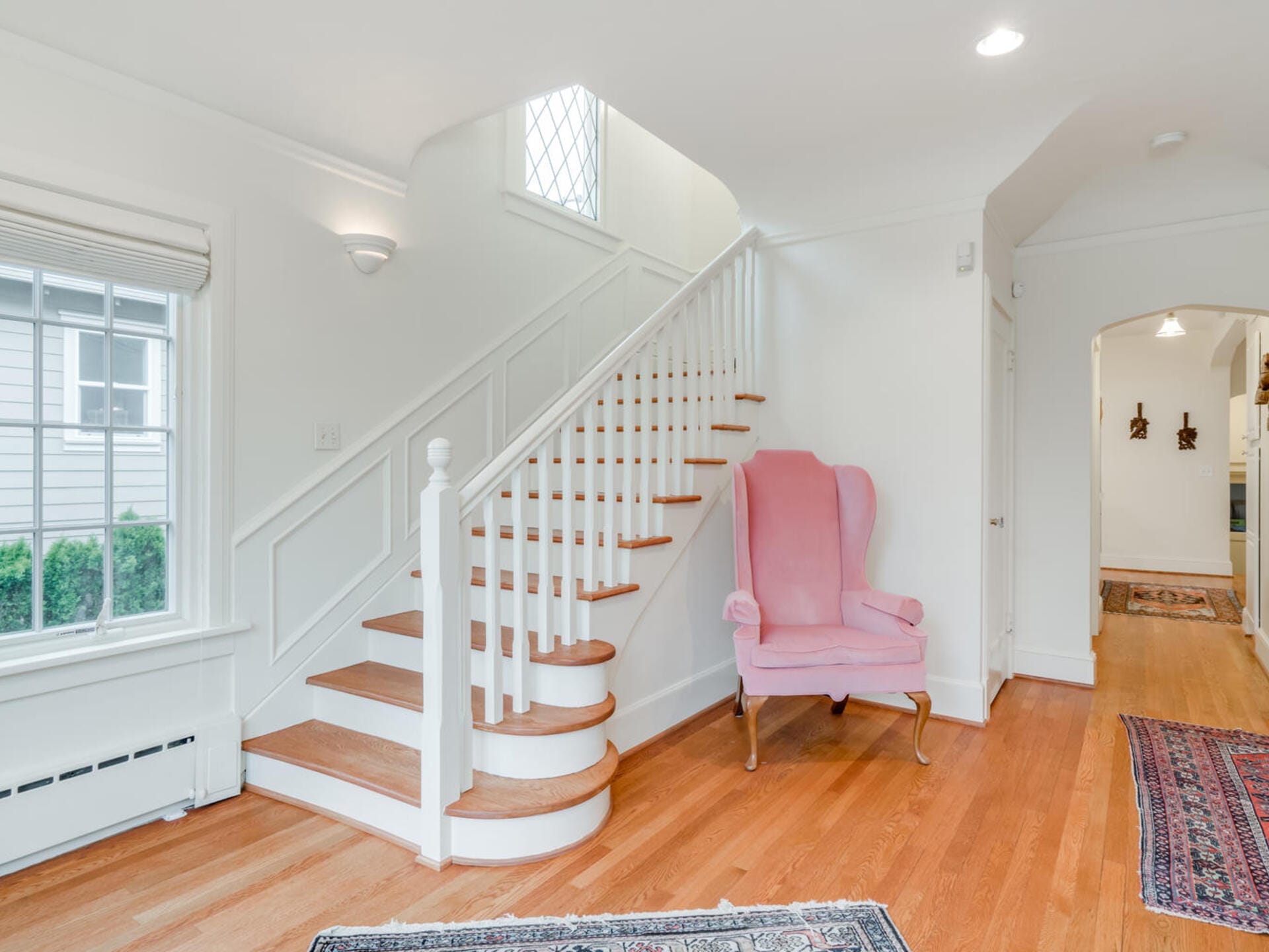 A bright hallway with a wooden staircase leading upstairs. A pink upholstered wingback chair sits by the railing. Light streams in through a window, illuminating the hardwood floor and a patterned rug. An archway leads to another room.