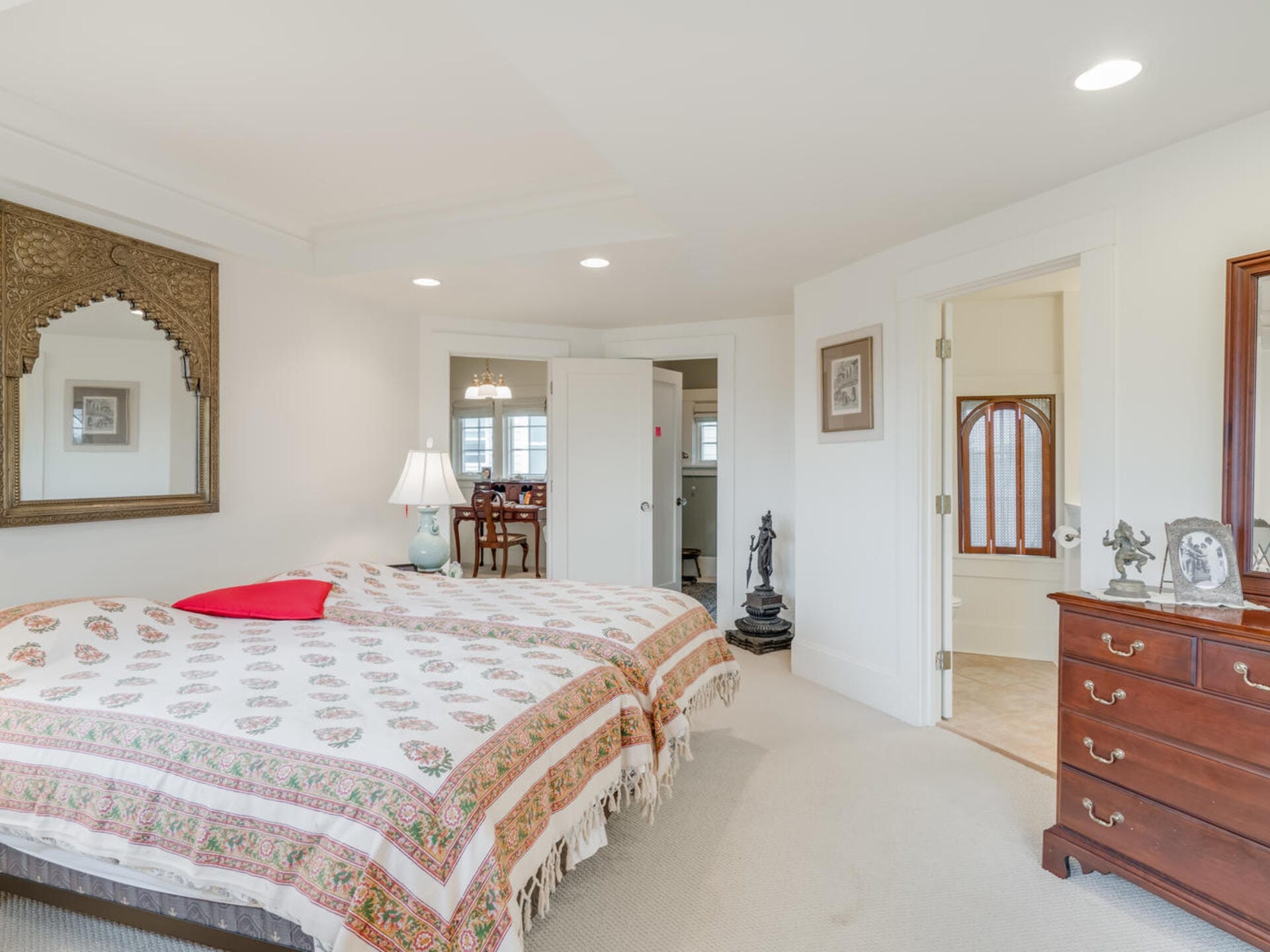 A cozy bedroom featuring a double bed with a patterned bedspread and a red pillow. A lamp on a side table provides soft lighting. Decorative wall art and a mirror adorn the walls. A doorway leads to an adjoining room and bathroom.